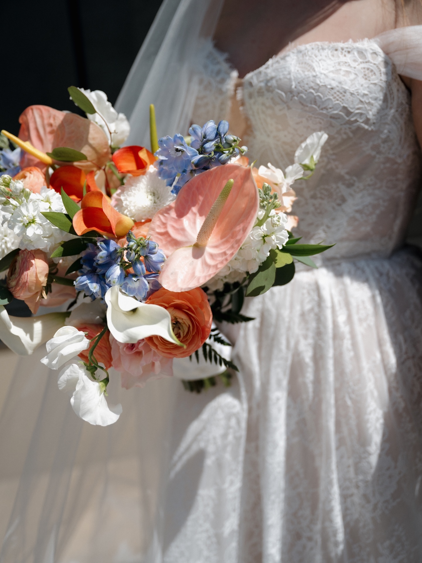 A vibrant close-up of the summer wedding bridal bouquet featuring pink anthuriums, orange ranunculus, blue delphinium, white calla lilies, and sweet peas
