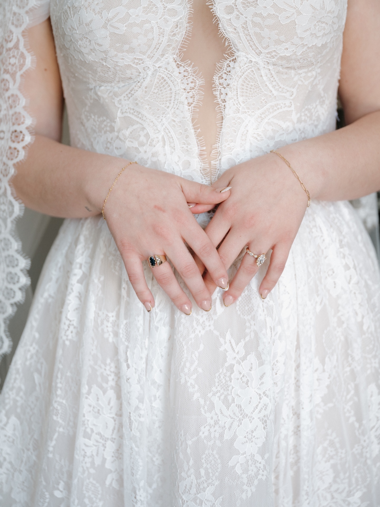 A close-up detail shot of the bride's hands resting against her lace gown, showing her engagement ring, wedding band, and delicate gold bracelets
