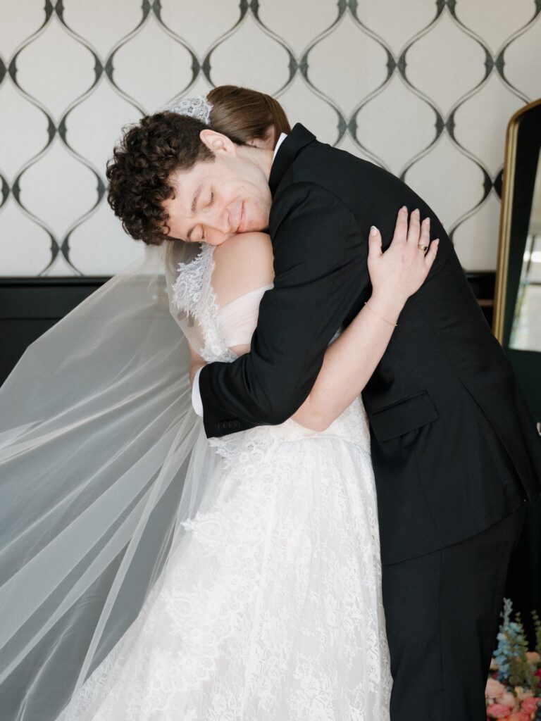 The groom buries his face in the bride's neck during an emotional first look embrace, her lace cathedral veil sweeping behind them
