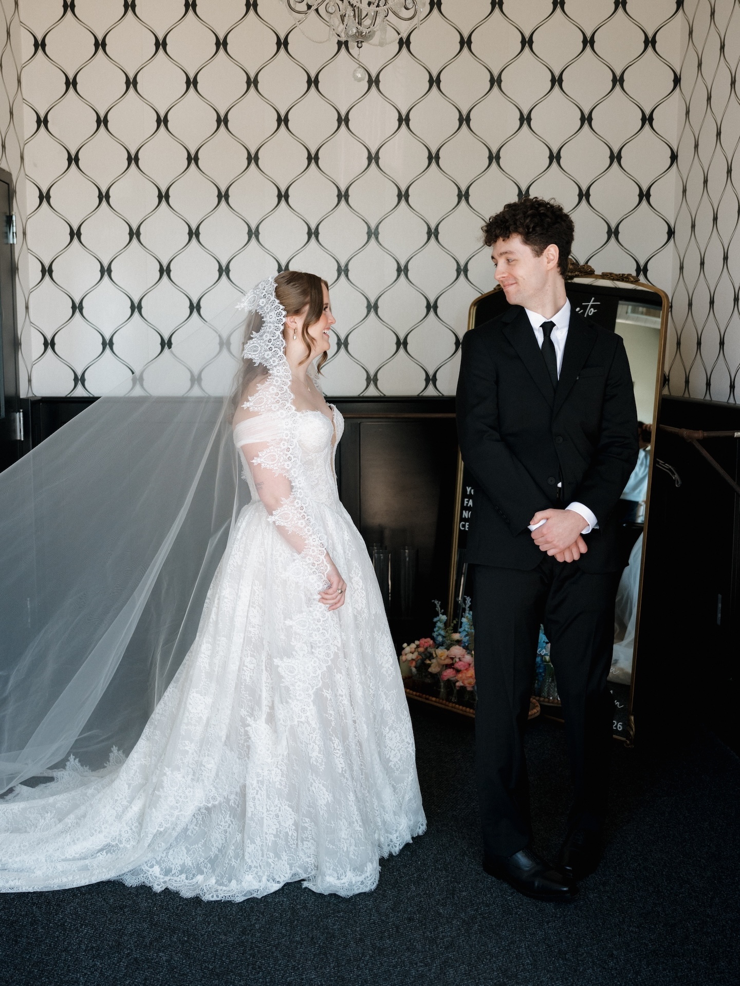 A summer wedding first look moment as the groom smiles at his bride in her lace gown and cathedral veil against a geometric wallpapered backdrop
