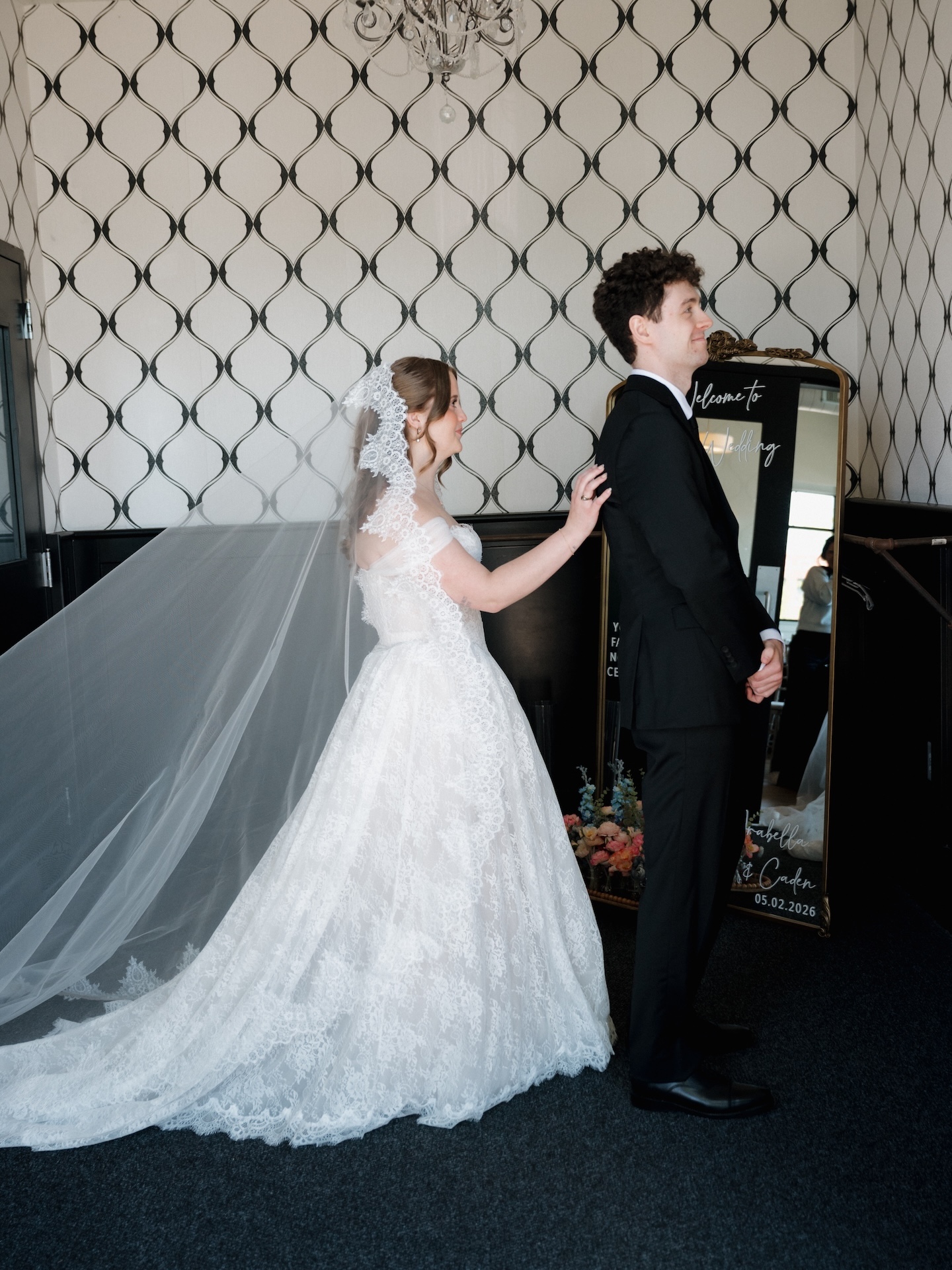 A bride in a lace ball gown and mantilla veil stands beside the groom in front of a welcome mirror sign reading "Arabella & Caden 05.02.2026"
