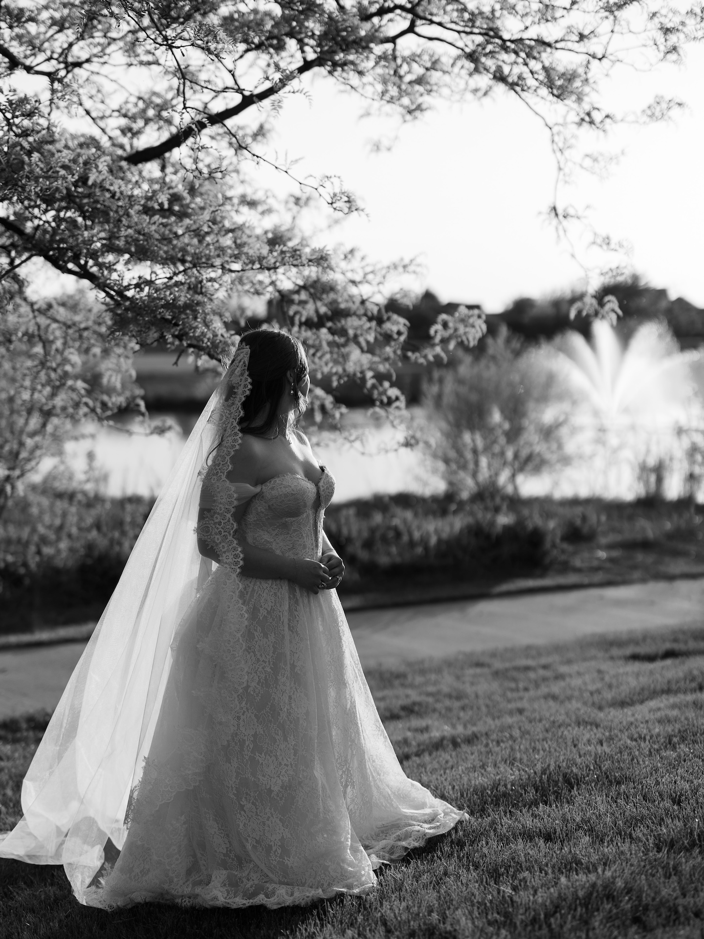 A summer wedding bridal portrait from behind as the bride in her lace ball gown and mantilla veil gazes toward a fountain amid golden hour light
