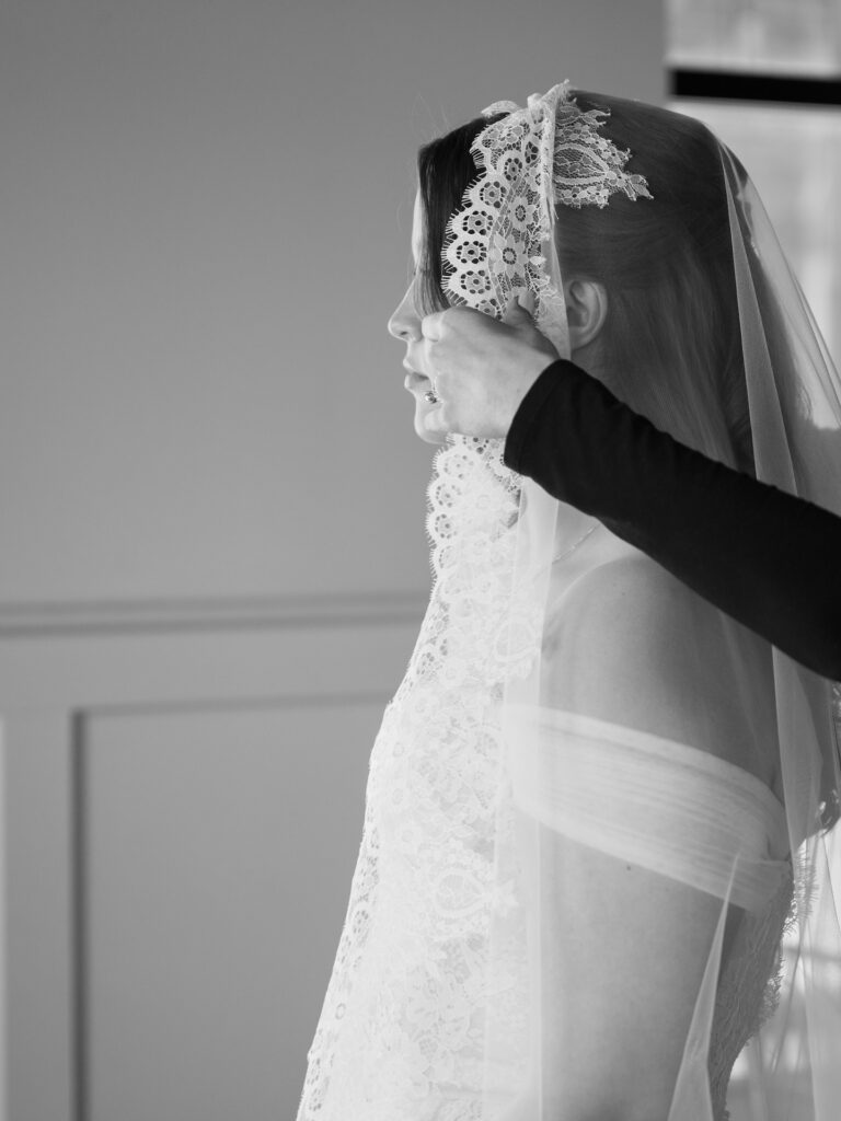 A black and white close-up profile of the bride as someone carefully pins her lace mantilla veil into her hair during summer wedding preparations
