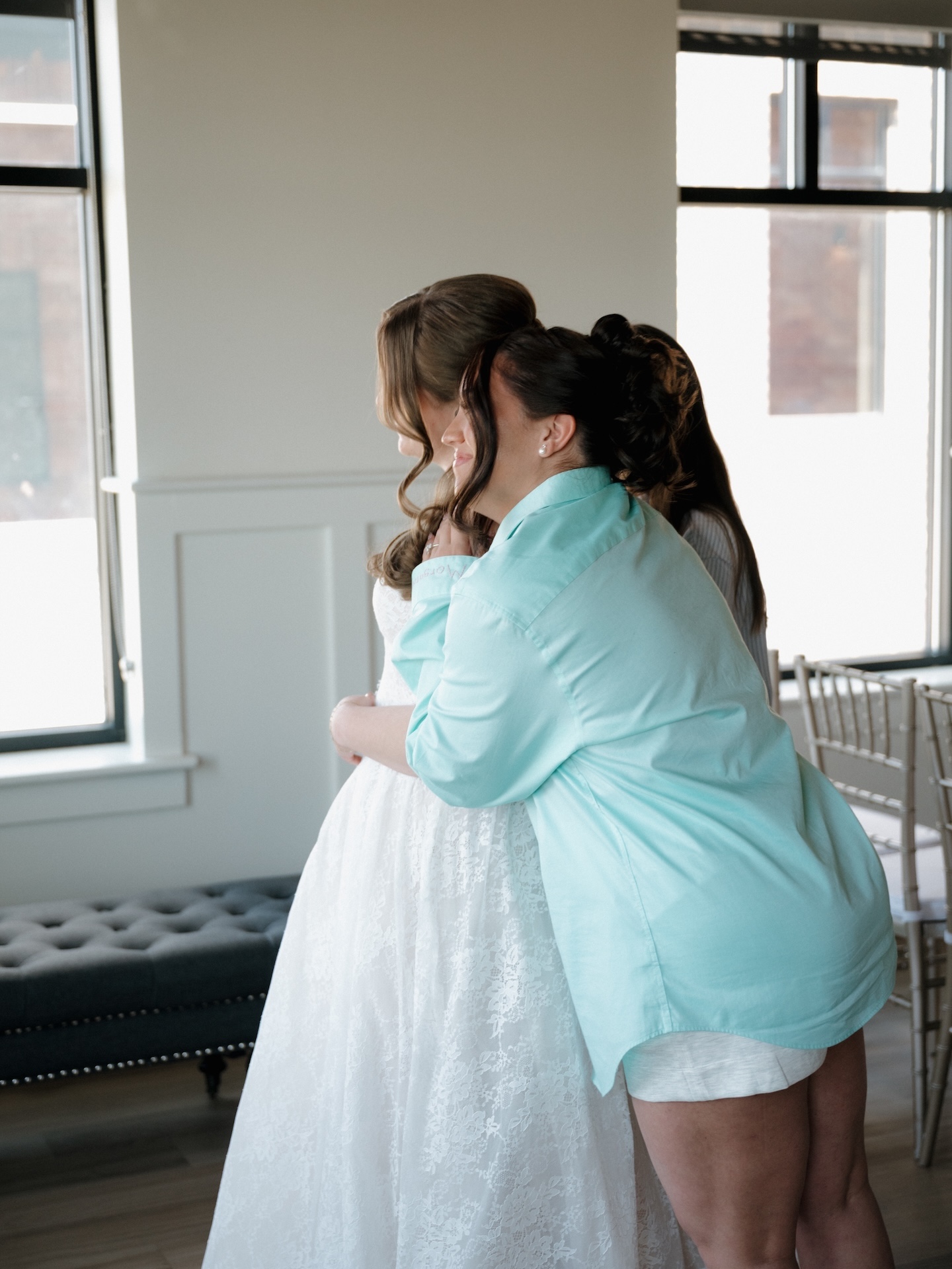 A summer wedding bridesmaid in a teal shirt embraces the bride in her lace gown near a bright window
