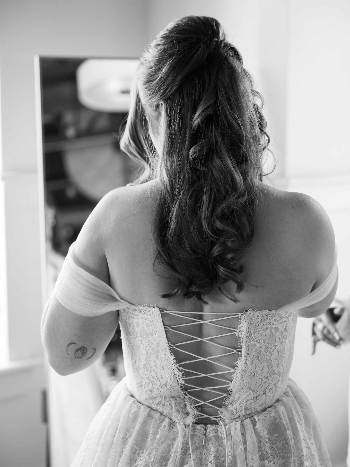 A black and white detail shot of the bride's lace corset gown from behind, showing the ribbon lace-up back and her half-up curled hair
