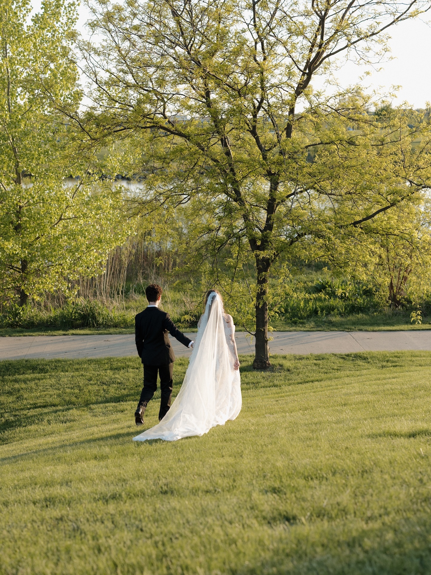 The newlyweds walk hand in hand across a lush green lawn toward a tree-lined lake during their summer wedding portrait session
