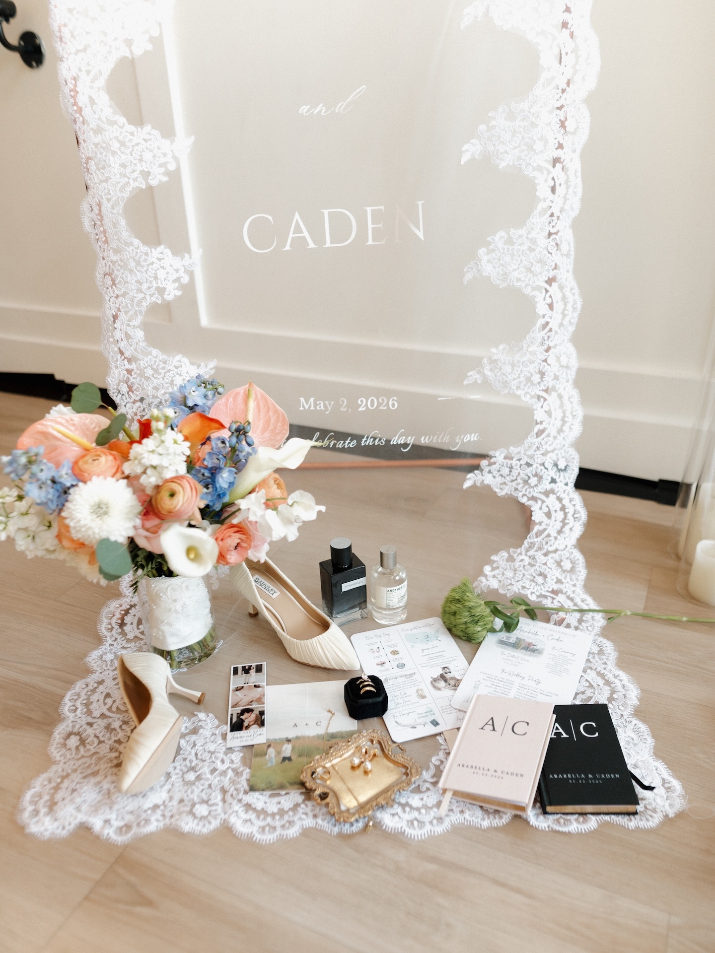A summer wedding flat lay styled around an acrylic welcome sign reading "Arabella and Caden, May 2, 2026," surrounded by shoes, florals, rings, and stationery
