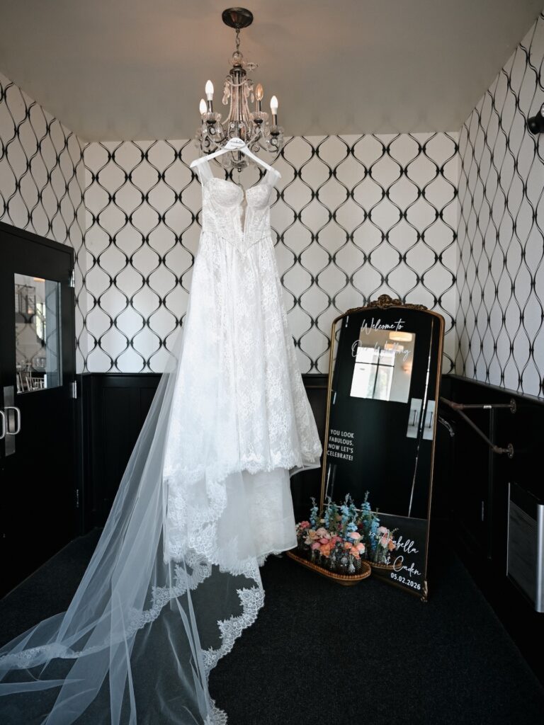 The lace bridal gown and cathedral veil hang from a chandelier beside the welcome mirror sign reading "Arabella & Caden 05.02.2026" in the bridal suite
