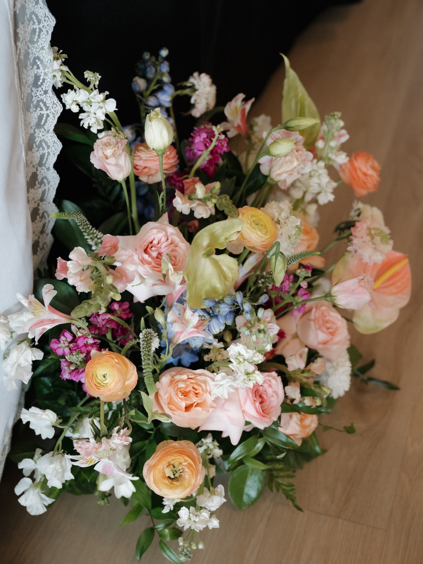 A close-up overhead shot of the bridal bouquet featuring peach garden roses, orange ranunculus, blue delphinium, green anthuriums, and magenta blooms
