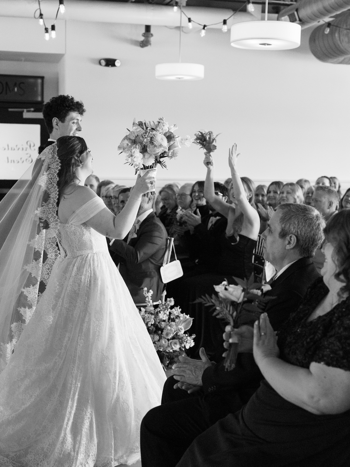 A black and white photo of the newlyweds walking back up the aisle as guests cheer and the bride raises her bouquet in celebration
