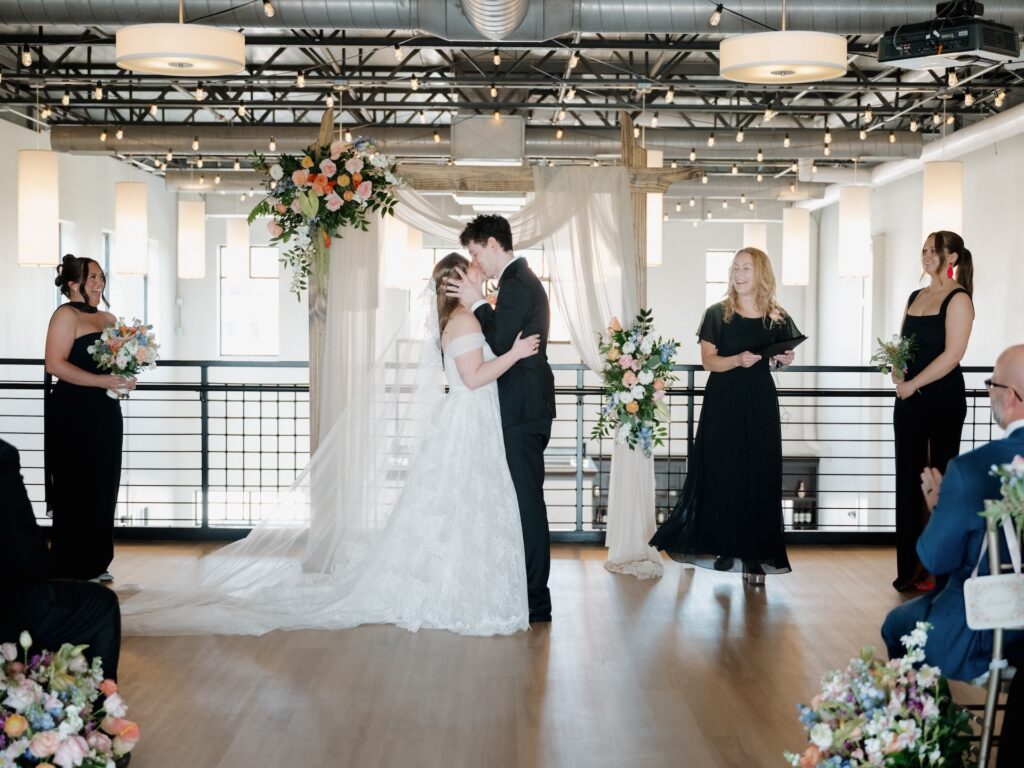 The bride and groom share their first kiss as a married couple beneath a floral arch at their summer wedding ceremony at The District in Des Moines
