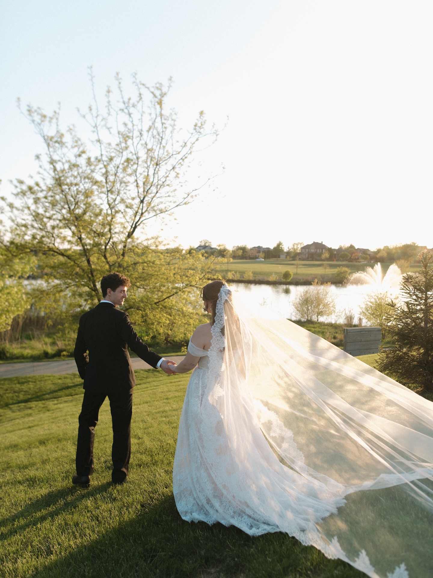 A summer wedding golden hour portrait as the couple walks hand in hand across a green lawn, the lace cathedral veil billowing in the warm light beside a fountain
