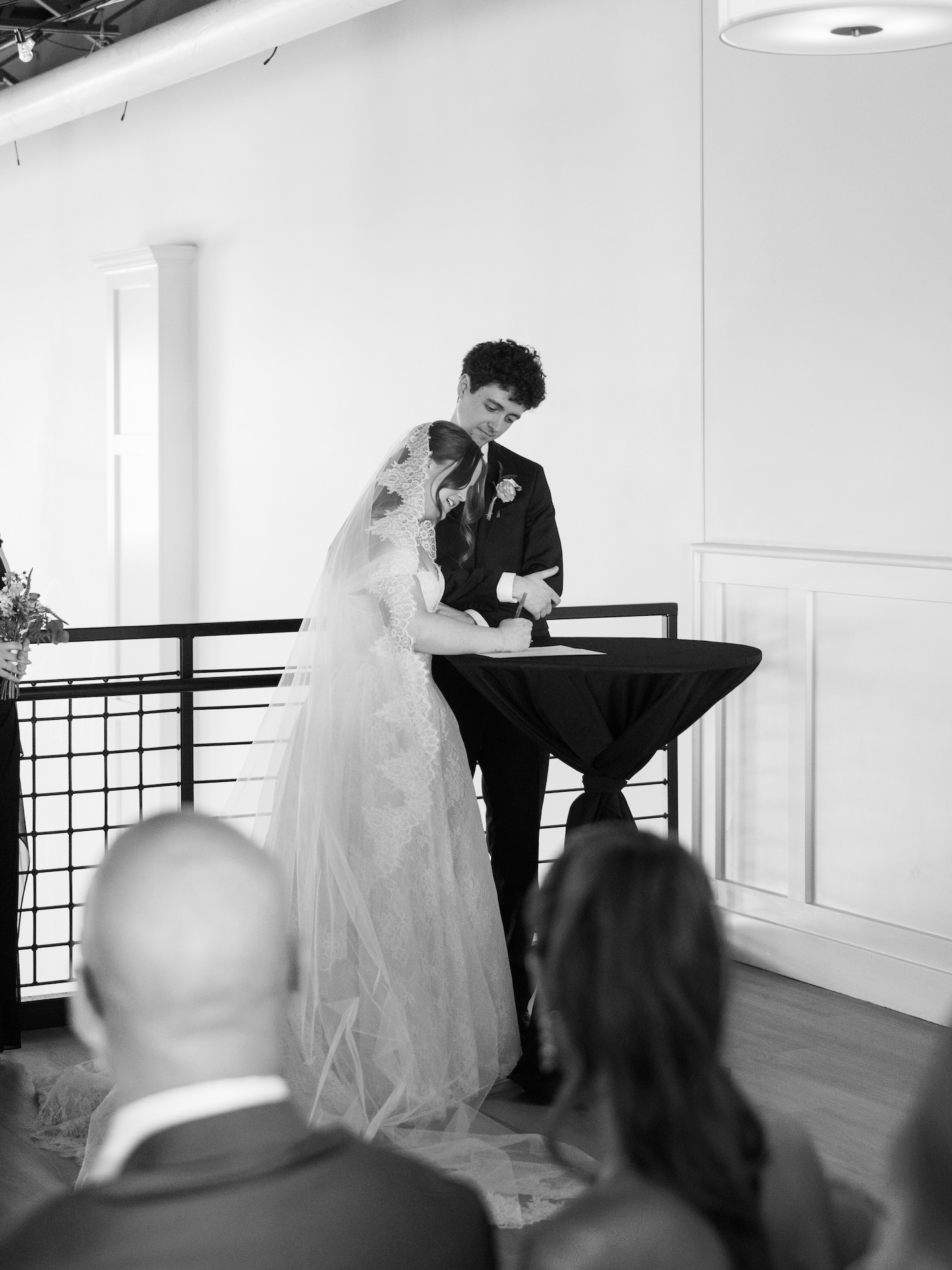A black and white photo of the bride and groom signing their marriage license at a draped cocktail table during the summer wedding ceremony at The District
