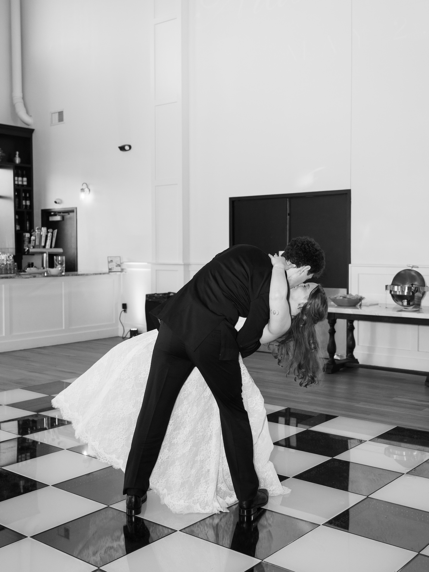 A black and white photo of the groom dipping the bride for a dramatic kiss on the checkered dance floor during their first dance
