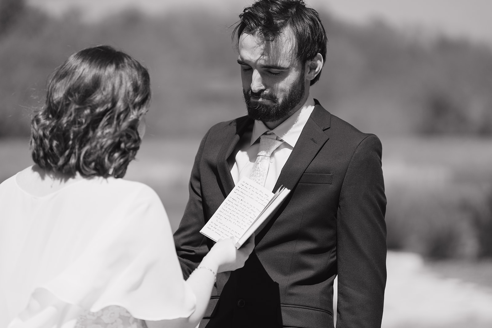 Black and white image of groom listening intently as his bride reads her handwritten vows during their outdoor ceremony