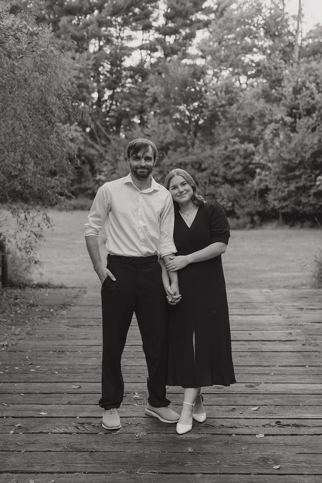 Fall engagement photos of a newly engaged couple standing arm in arm on a wooden footbridge smiling at the camera in black and white
