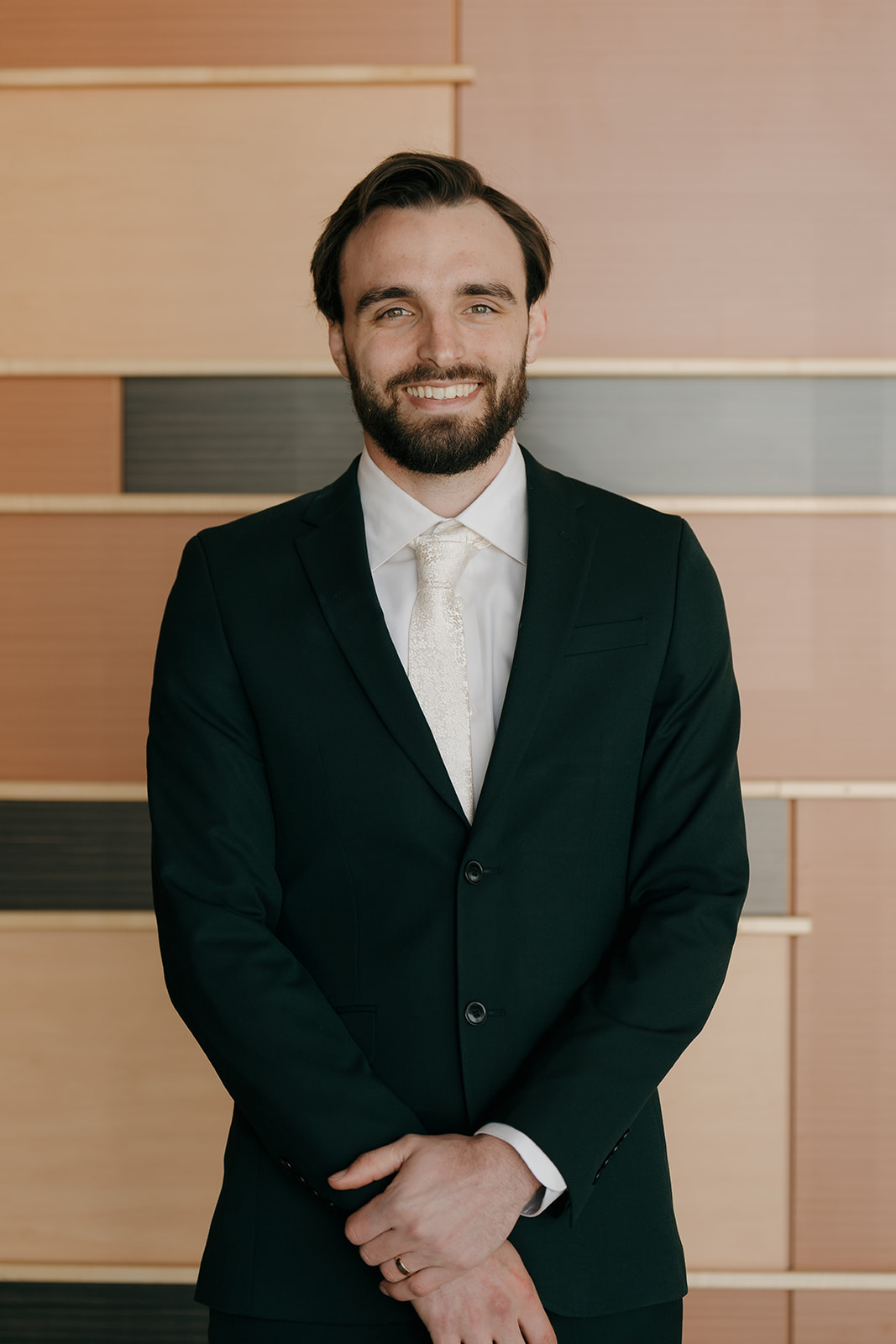 Groom smiling confidently in a dark suit and cream tie against a warm-toned paneled wall, captured in wedding photography styles that blend elegance and personality