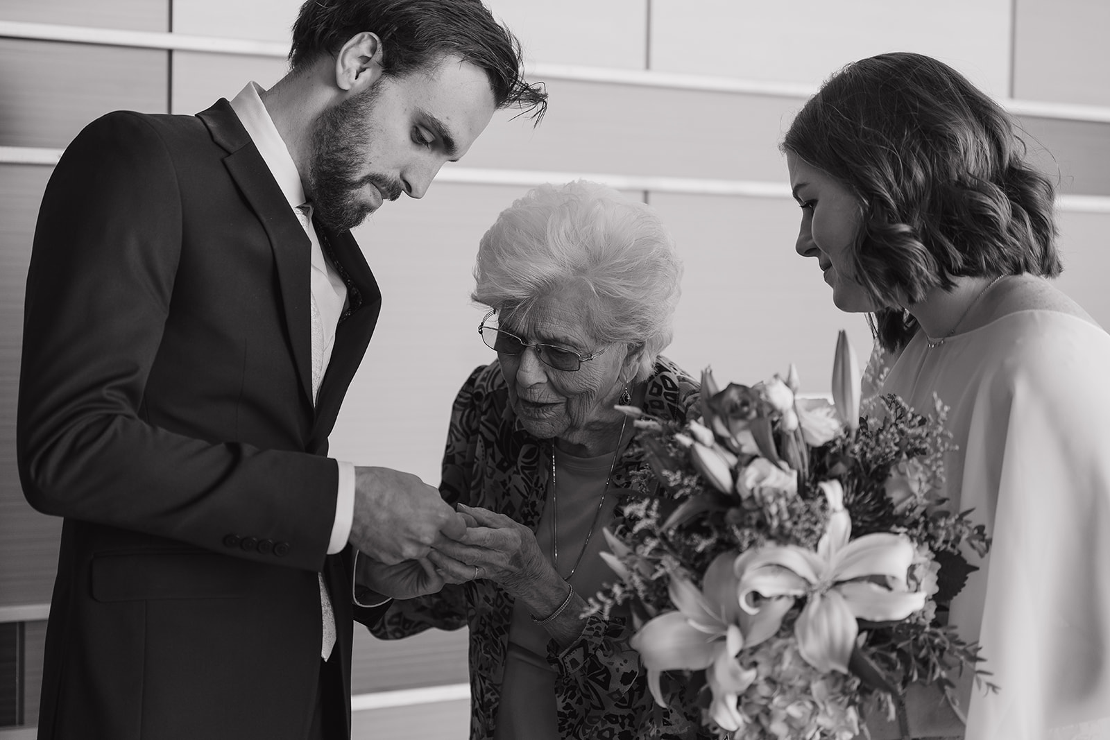 Black and white photo of bride and groom showing their wedding rings to an elderly family member after the ceremony, captured in wedding photography styles that honor family moments