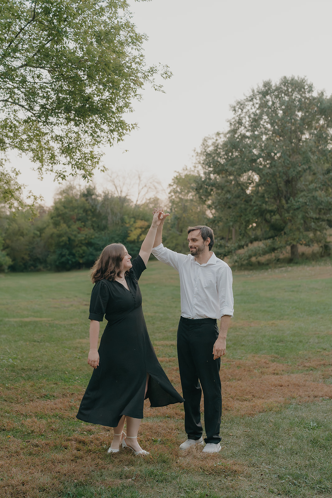 Fall engagement photos of a couple dancing together in an open park field at golden hour, her dress swirling as he spins her
