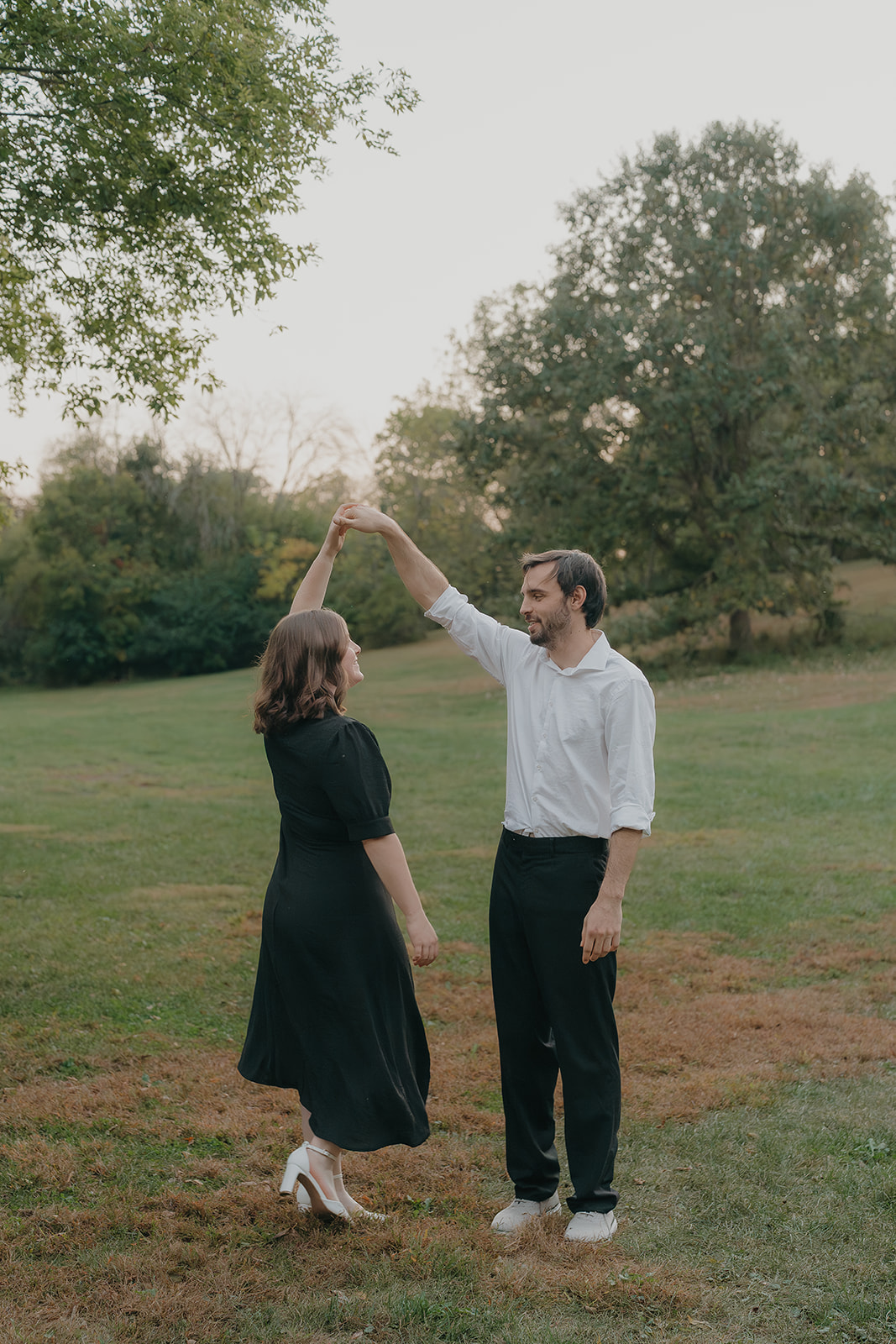 Fall engagement photos of a couple dancing in a grassy park field at golden hour, him spinning her under his arm