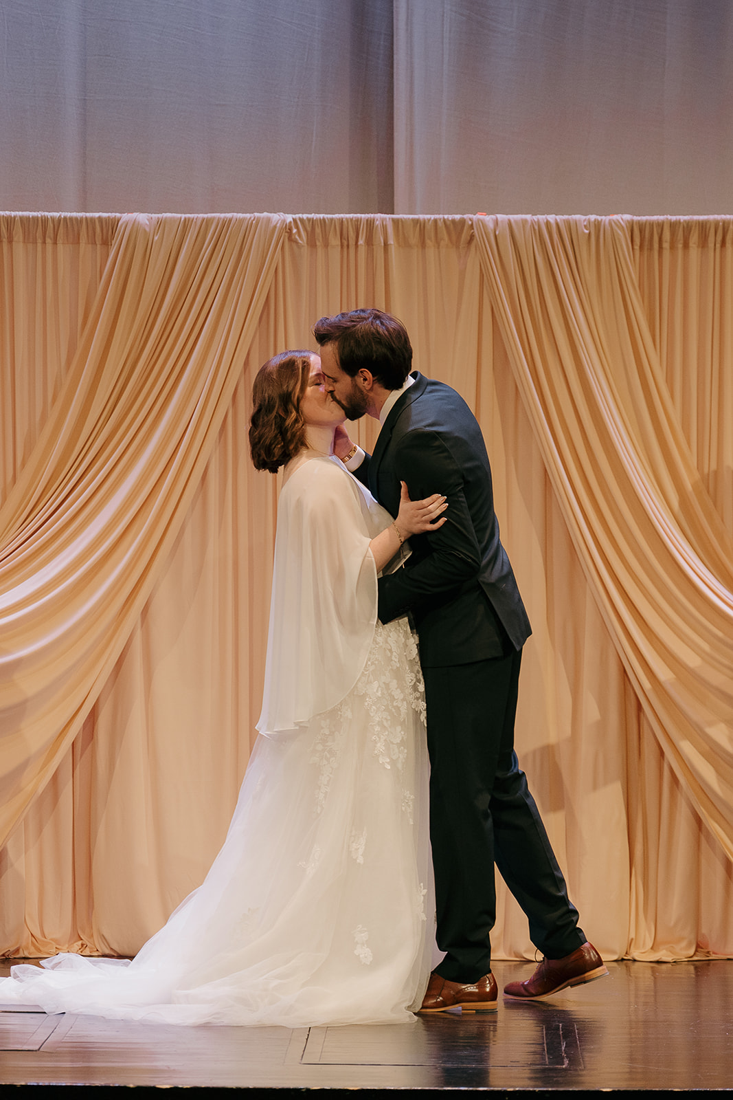 Bride and groom sharing their first kiss in front of a draped blush backdrop, captured in wedding photography styles that blend elegance and emotion