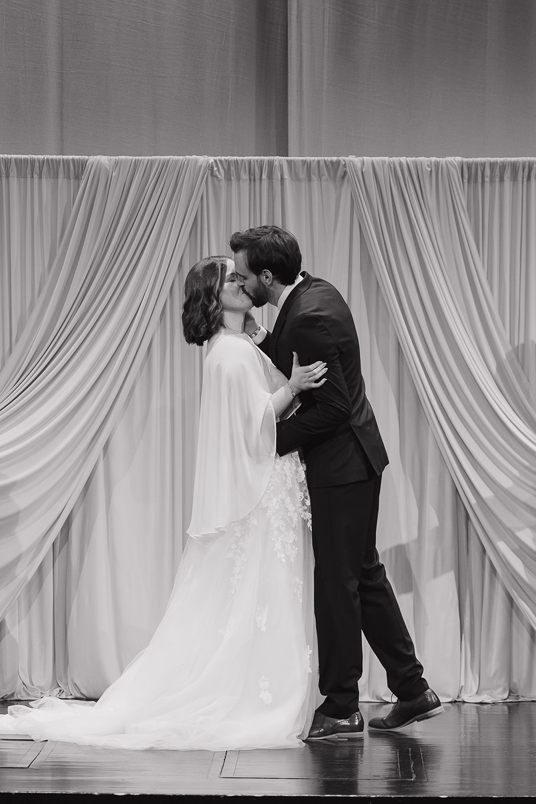 Black and white image of bride and groom sharing their first kiss in front of a draped ceremony backdrop