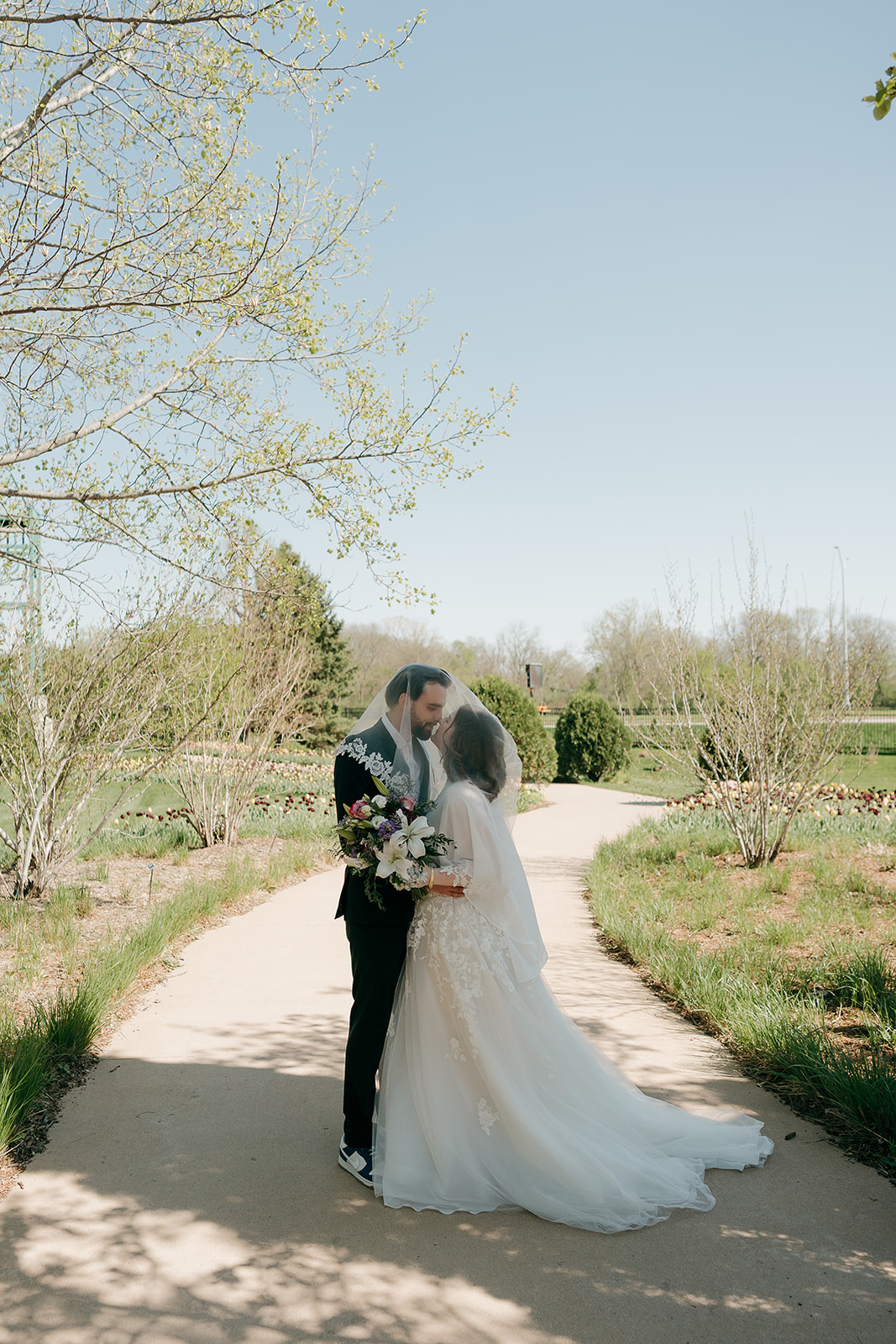 Bride and groom kissing under her lace-trimmed veil on a garden path surrounded by spring blooms