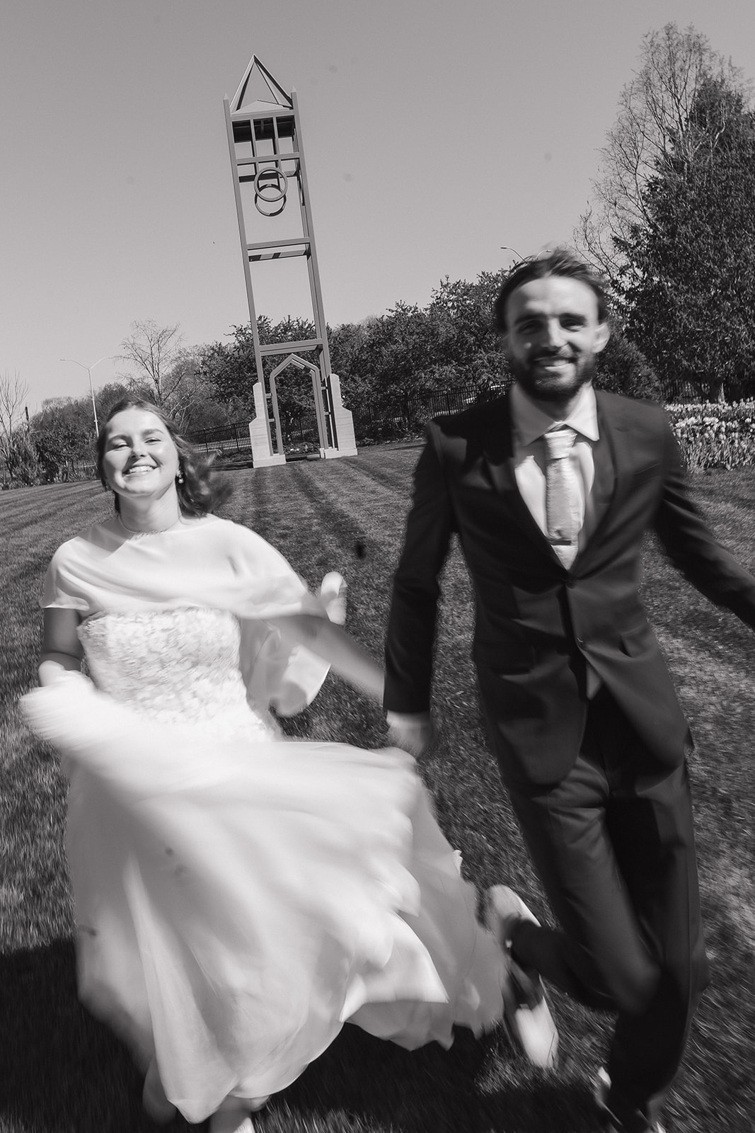 Joyful black and white photo of bride and groom running and laughing together in front of a bell tower, captured in wedding photography styles full of genuine happiness