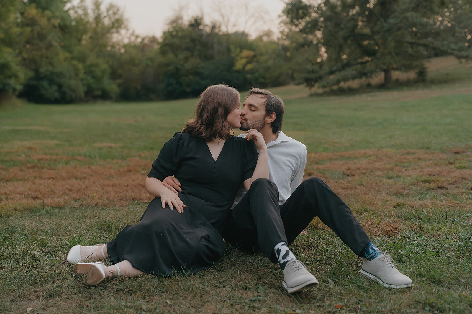 Fall engagement photos of a couple sitting in the grass kissing at dusk with a soft pink sky behind them