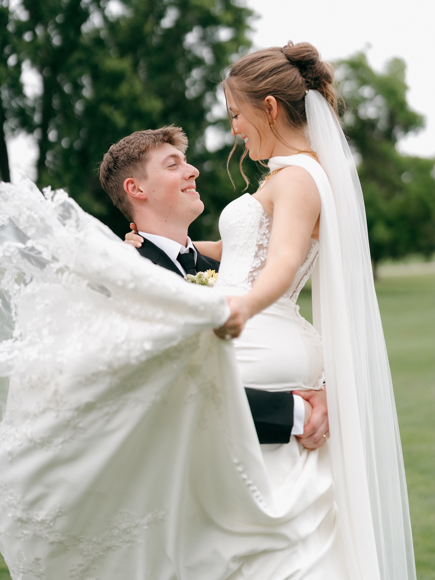Groom lifting his smiling bride on a golf course, her lace-appliqué wedding gown billowing as her veil flows behind her, yellow bridesmaid dresses worn by her bridal party