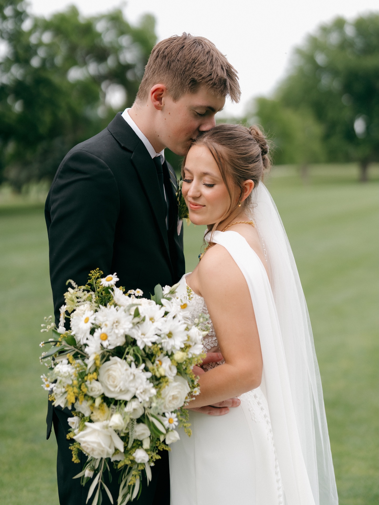 Groom pressing a kiss to the bride's forehead while she closes her eyes and smiles, holding a lush white and daisy bridal bouquet, from a wedding with yellow bridesmaid dresses
