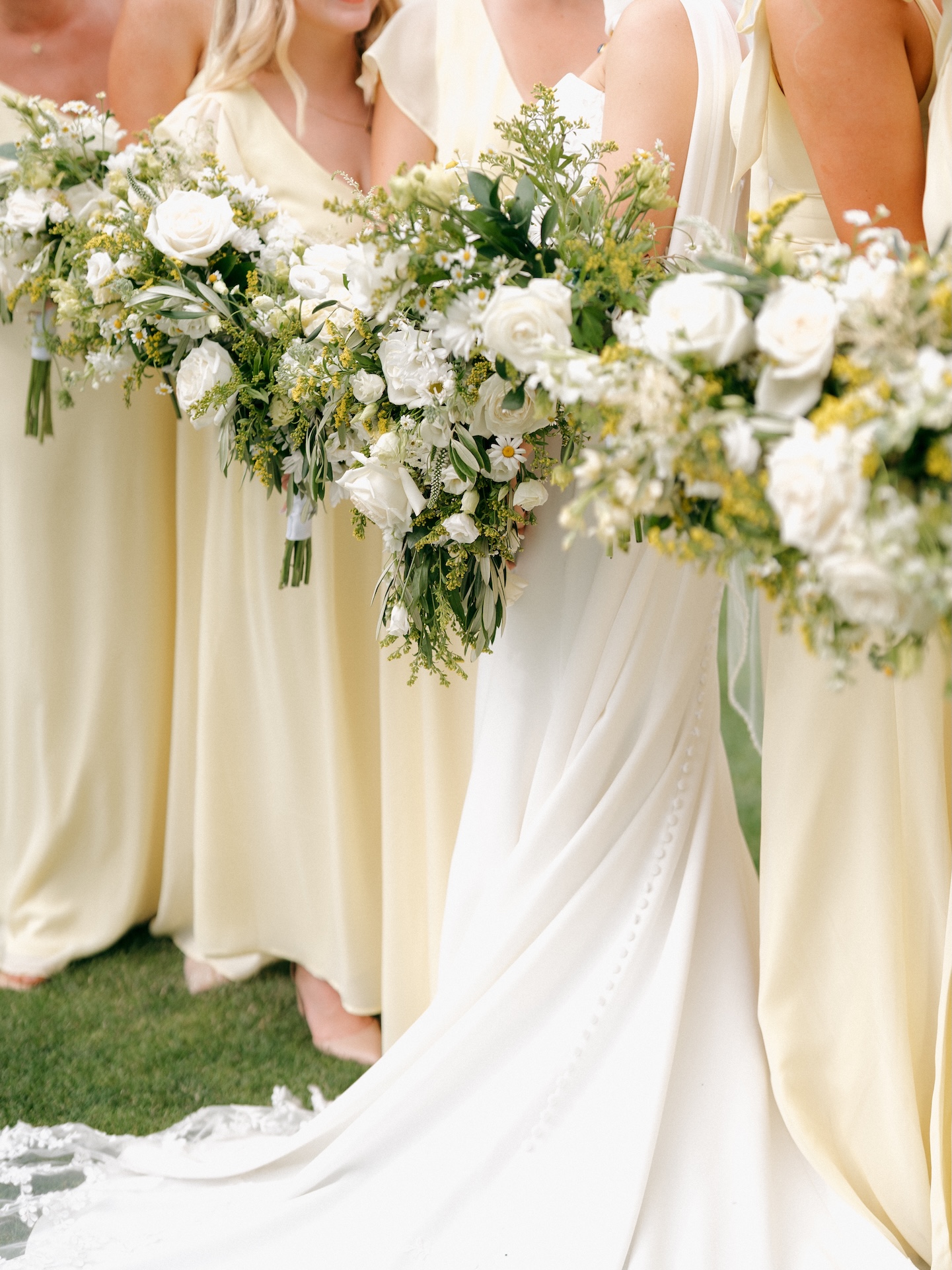 Close-up of bridesmaids in soft yellow bridesmaid dresses holding oversized white and greenery bouquets with yellow wildflower accents next to the bride in her white gown
