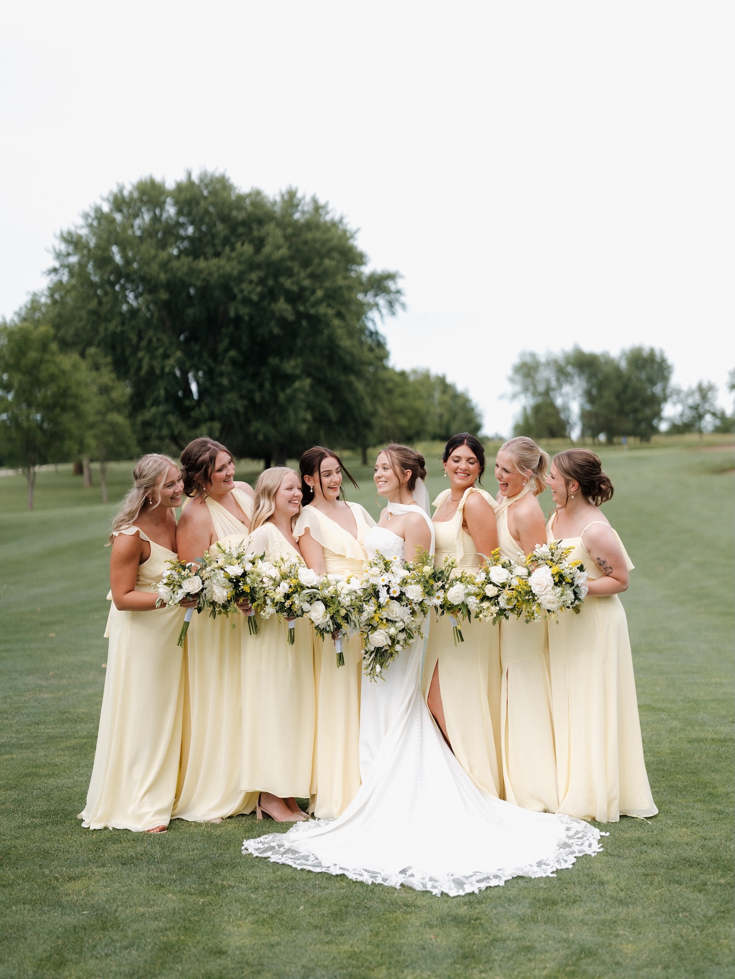 Bride surrounded by seven bridesmaids in soft yellow bridesmaid dresses on a golf course, all laughing together and holding white and greenery bouquets with pops of yellow
