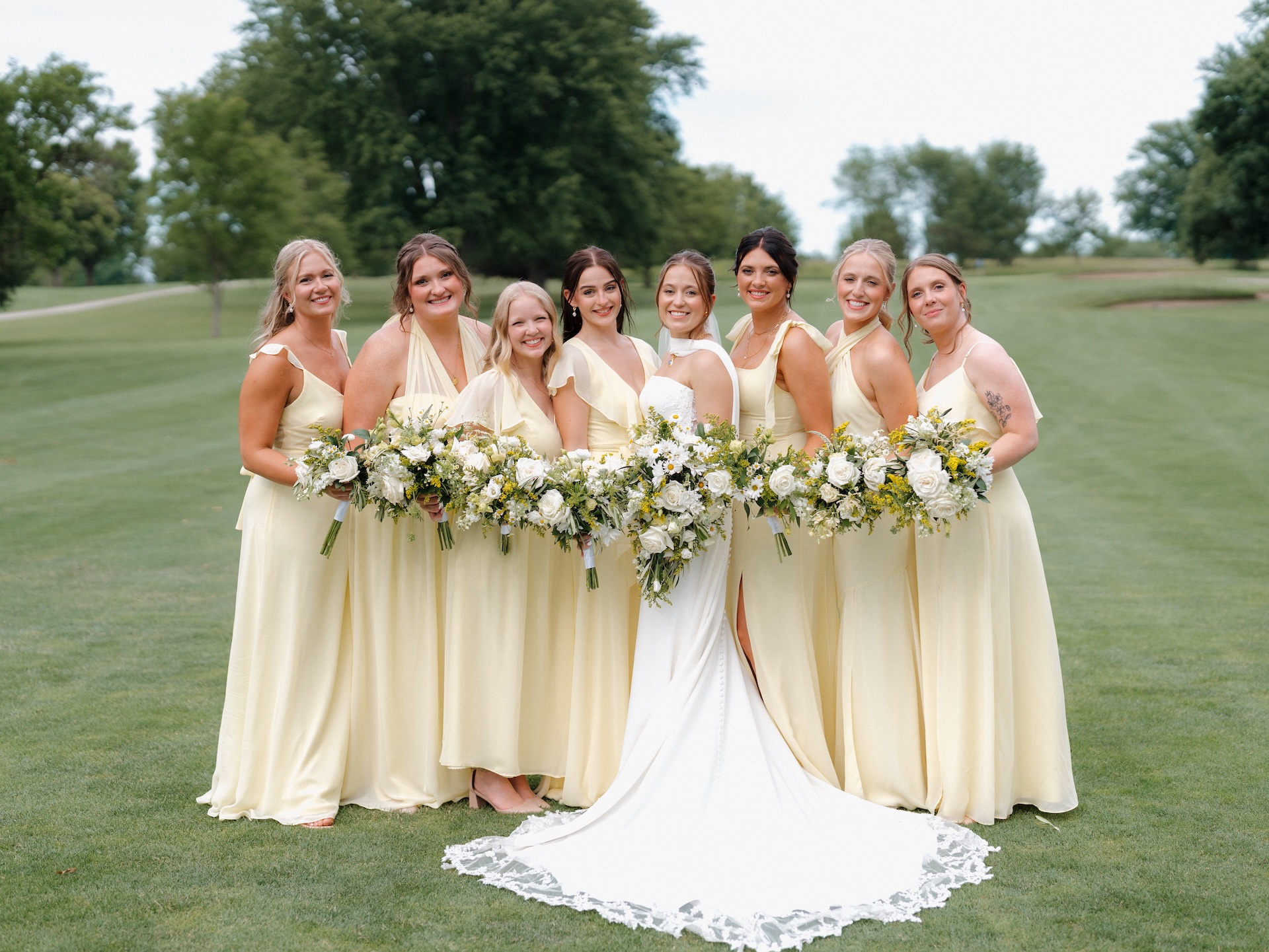 Bride standing at the center of her seven bridesmaids in yellow bridesmaid dresses on a golf course, all smiling at the camera and holding white and greenery bouquets with yellow wildflower accents
