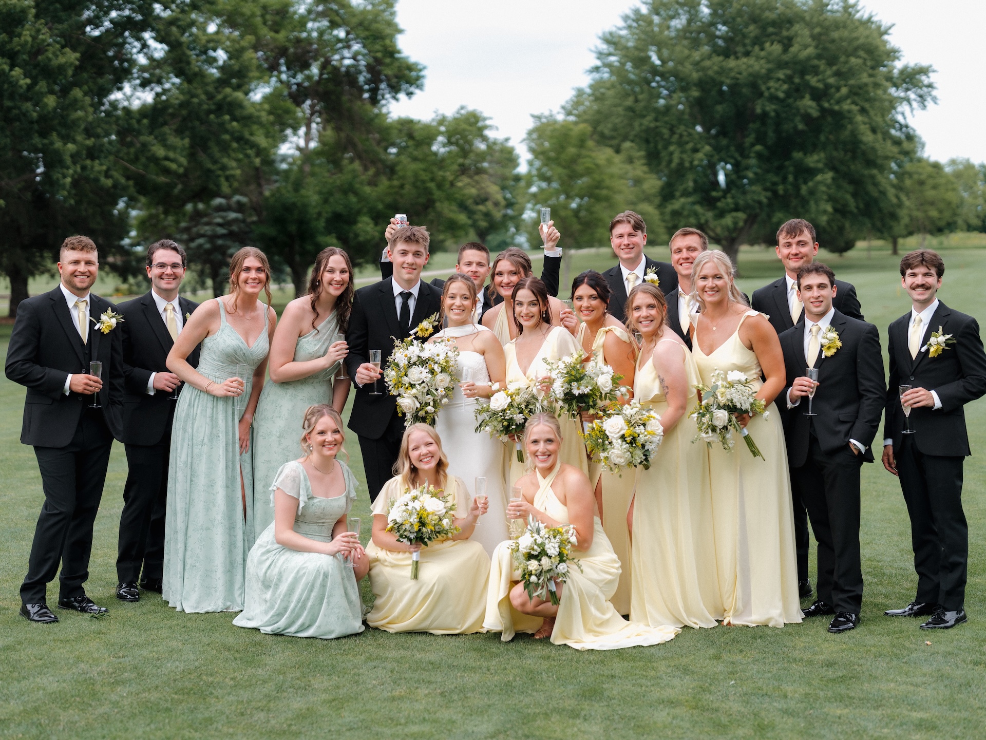 Full wedding party group photo on a golf course, bridesmaids in yellow bridesmaid dresses and sage green dresses mixed together, groomsmen in black suits with yellow ties, all raising champagne glasses
