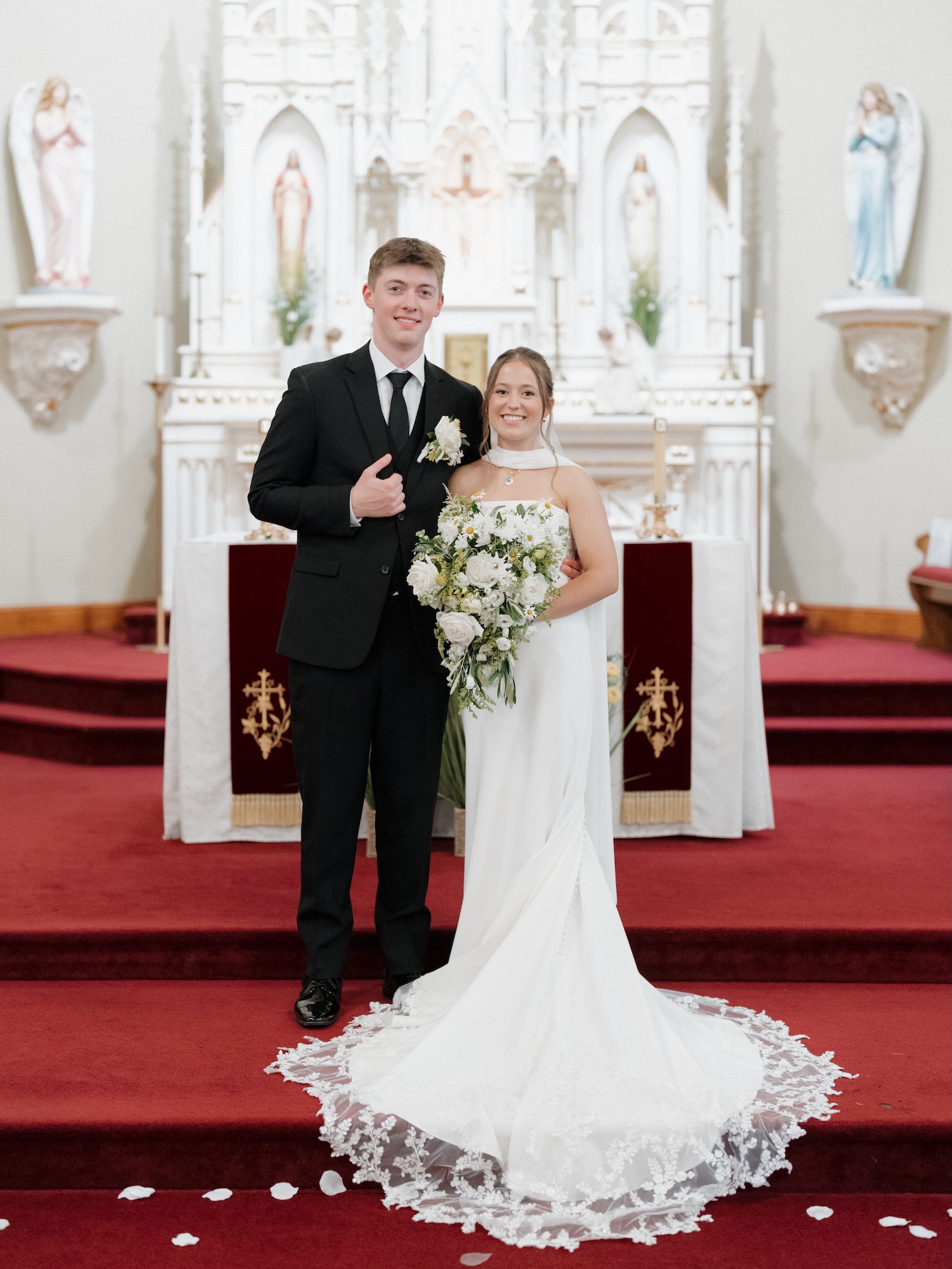 Bride and groom posing together at the altar of a Catholic church with a white ornate reredos and religious statues, the bride's lace-trimmed cathedral train spread across the red carpet, from a wedding with yellow bridesmaid dresses
