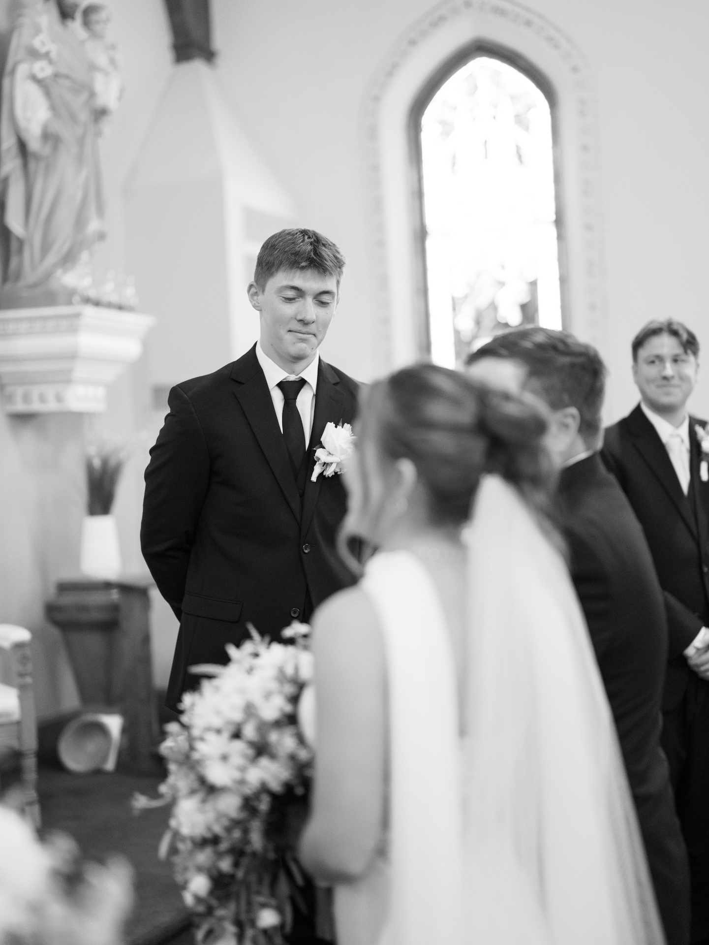 Black and white photo of the groom watching with an emotional expression as his bride walks toward him down the aisle inside a Catholic church, yellow bridesmaid dresses part of the wedding
