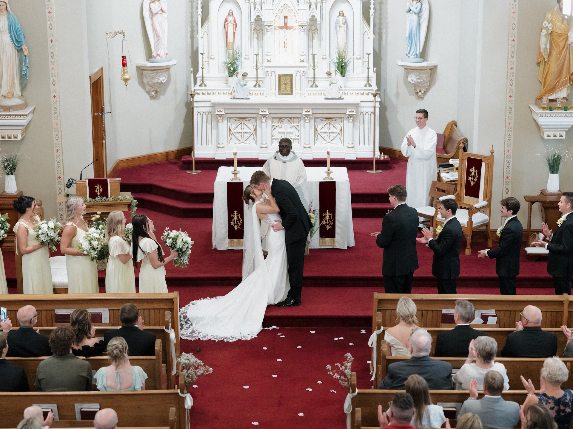 Wide shot of a first kiss at the altar inside a Catholic church, with yellow bridesmaid dresses lining the left side of the altar
