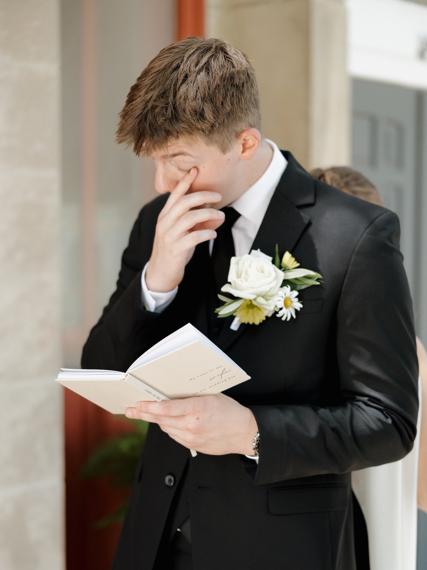 Groom in a black suit with a white and yellow boutonniere wiping tears while reading his vows from a small book labeled "My vows to my wife on our wedding day," yellow bridesmaid dresses worn by the bridal party
