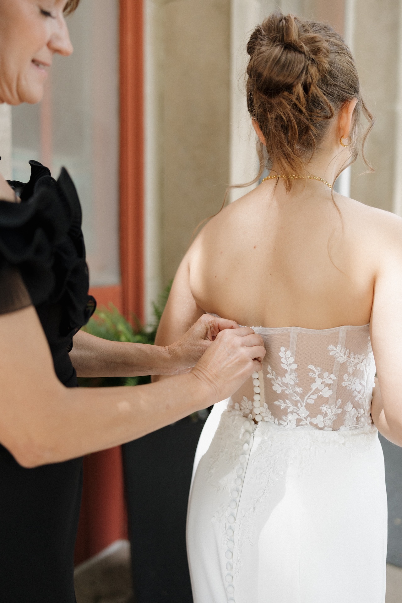 Mother buttoning the lace corset back of the bride's strapless wedding gown outside a brick church, from a wedding where the bridal party wore yellow bridesmaid dresses
