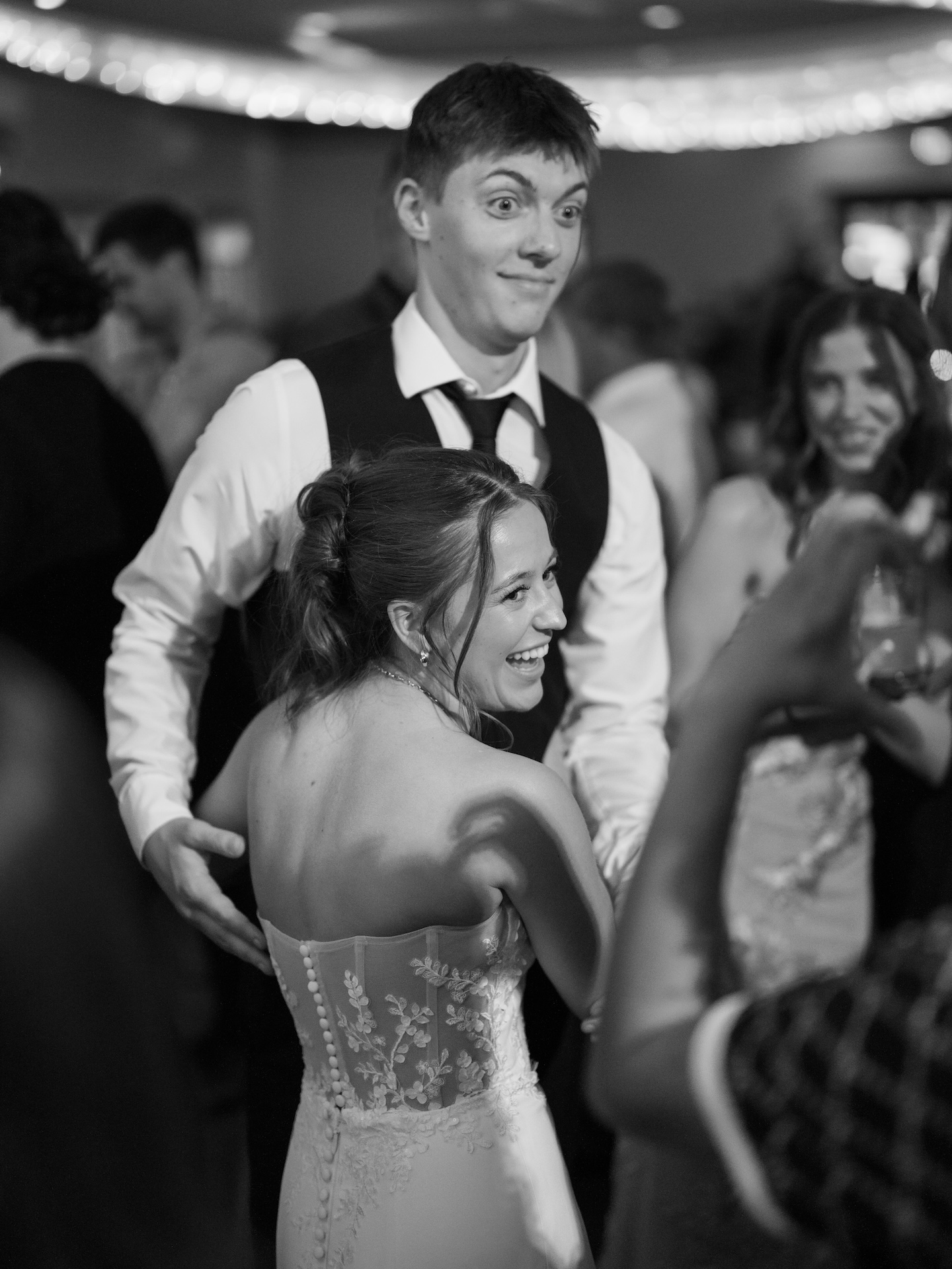 Black and white candid of bride and groom laughing and dancing together at their reception, the back of the bride's lace corset wedding gown visible, from a wedding with yellow bridesmaid dresses

