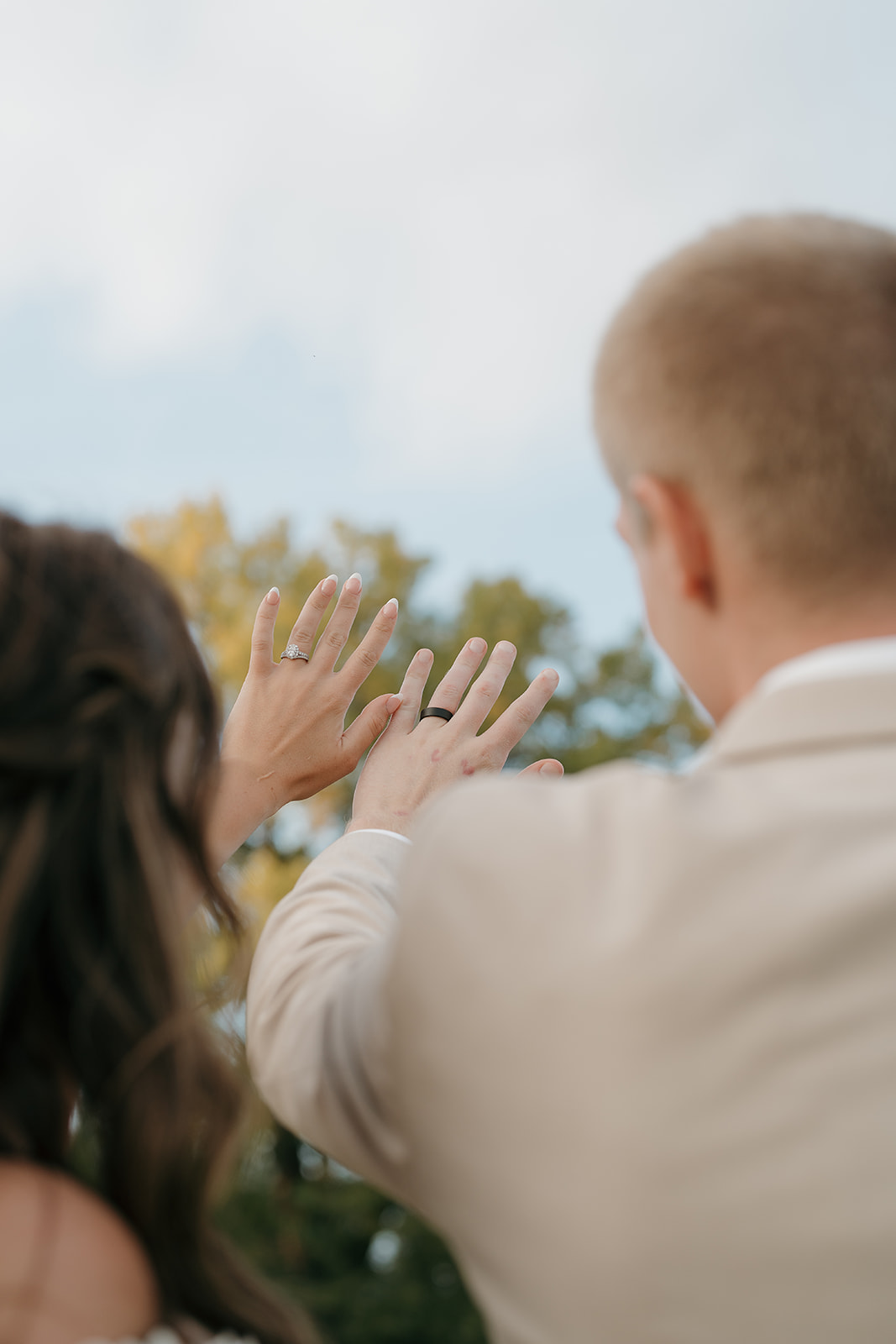Bride and groom raising their hands to show off wedding rings against a cloudy sky