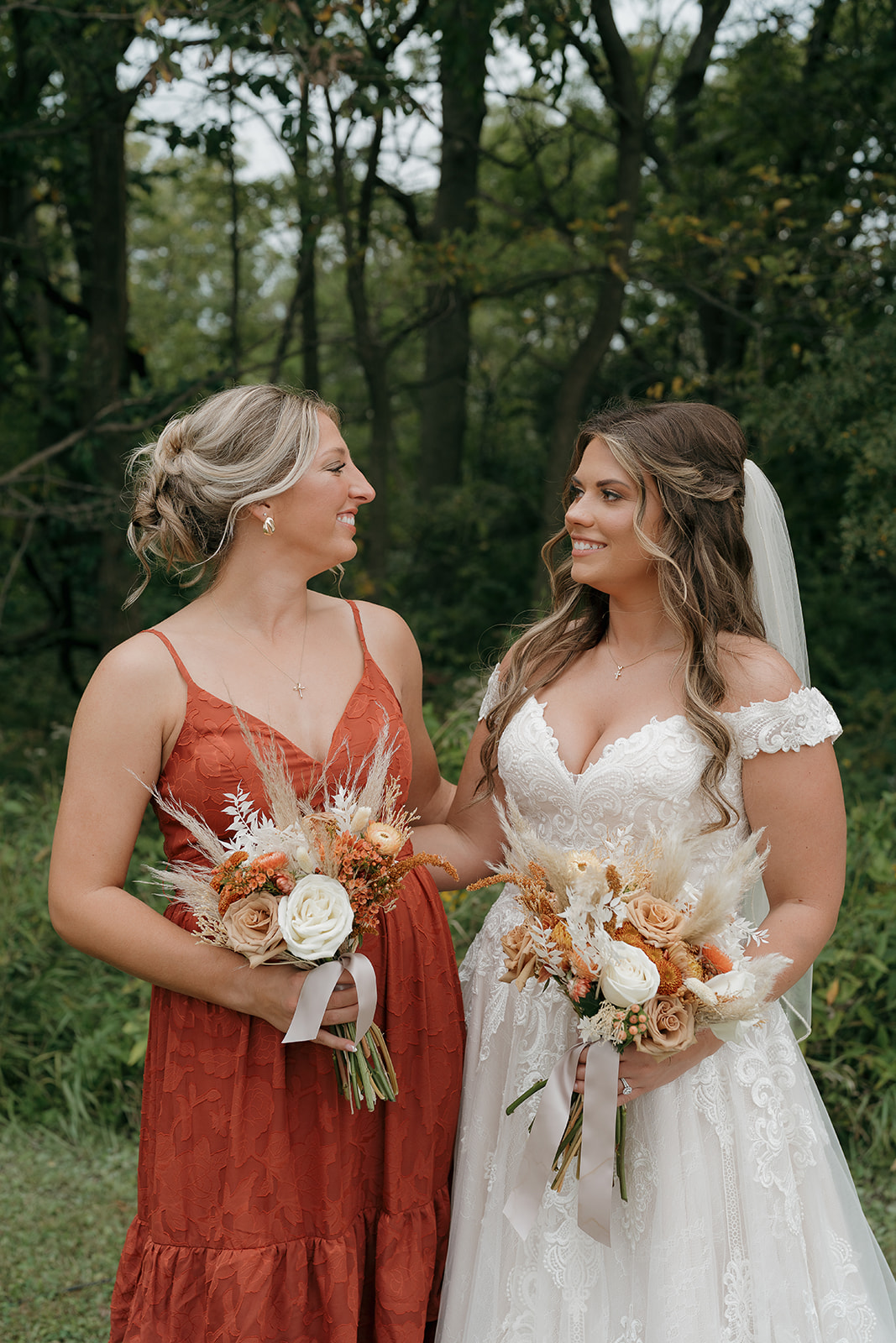 Bride and bridesmaid smiling at each other while holding matching bouquets in fall wedding colors