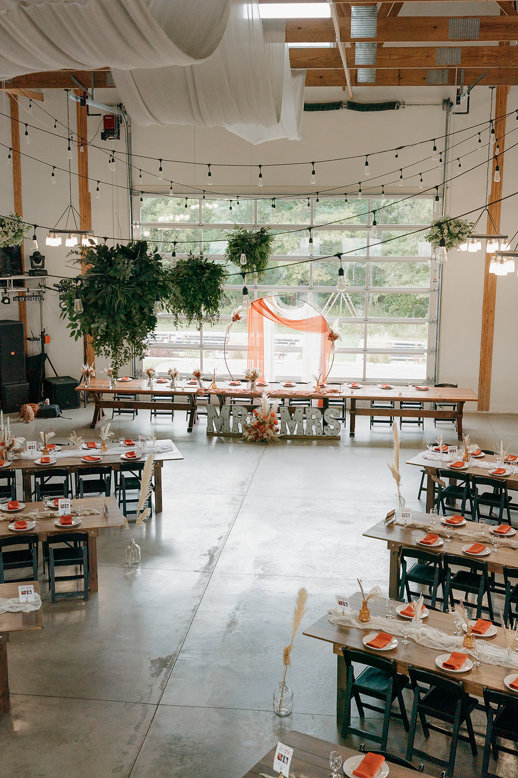 Reception hall set up with wooden farm tables, string lights, greenery, and an orange and white ceremony backdrop