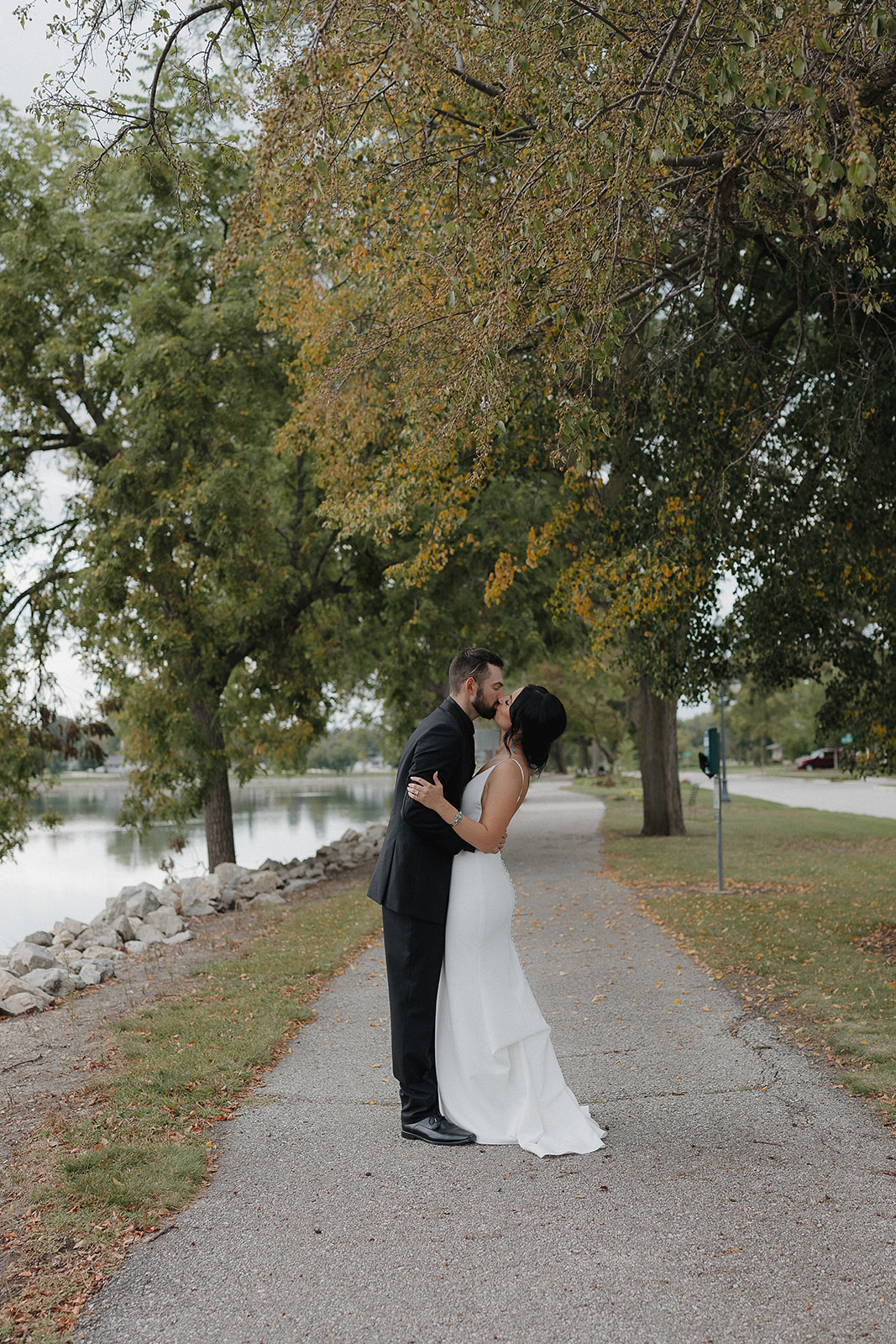 Bride and groom sharing a kiss on a tree lined path beside the lake