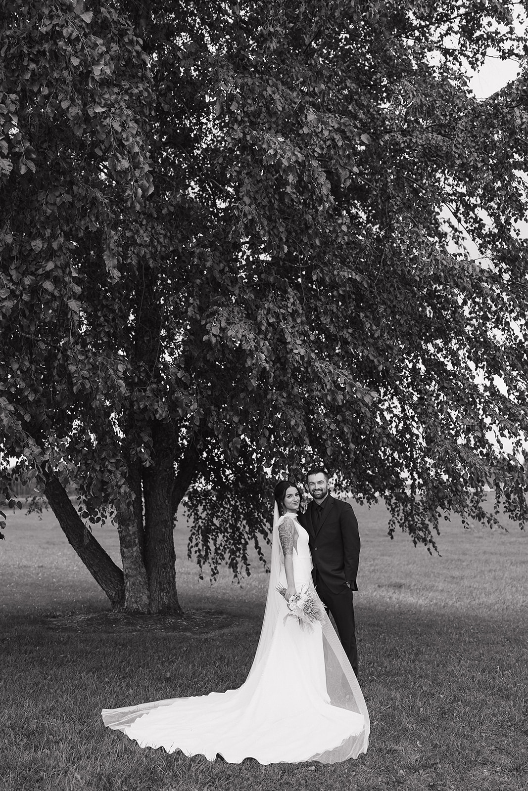 Black and white portrait of the couple standing together under a sprawling tree at their fall wedding