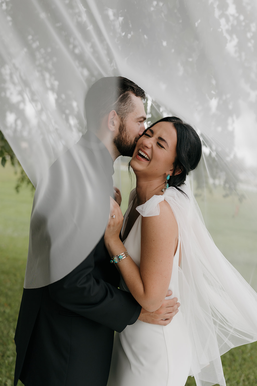 Veil wrapped portrait of the groom kissing the bride's cheek as she laughs, a romantic fall wedding moment