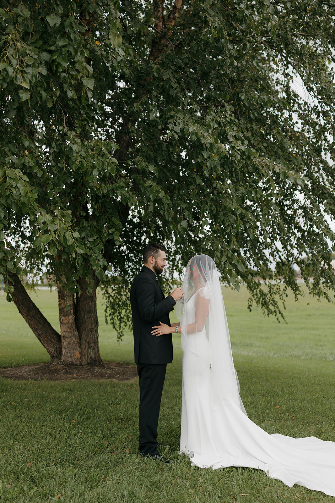 Groom helping the bride adjust her veil under a large tree before the ceremony