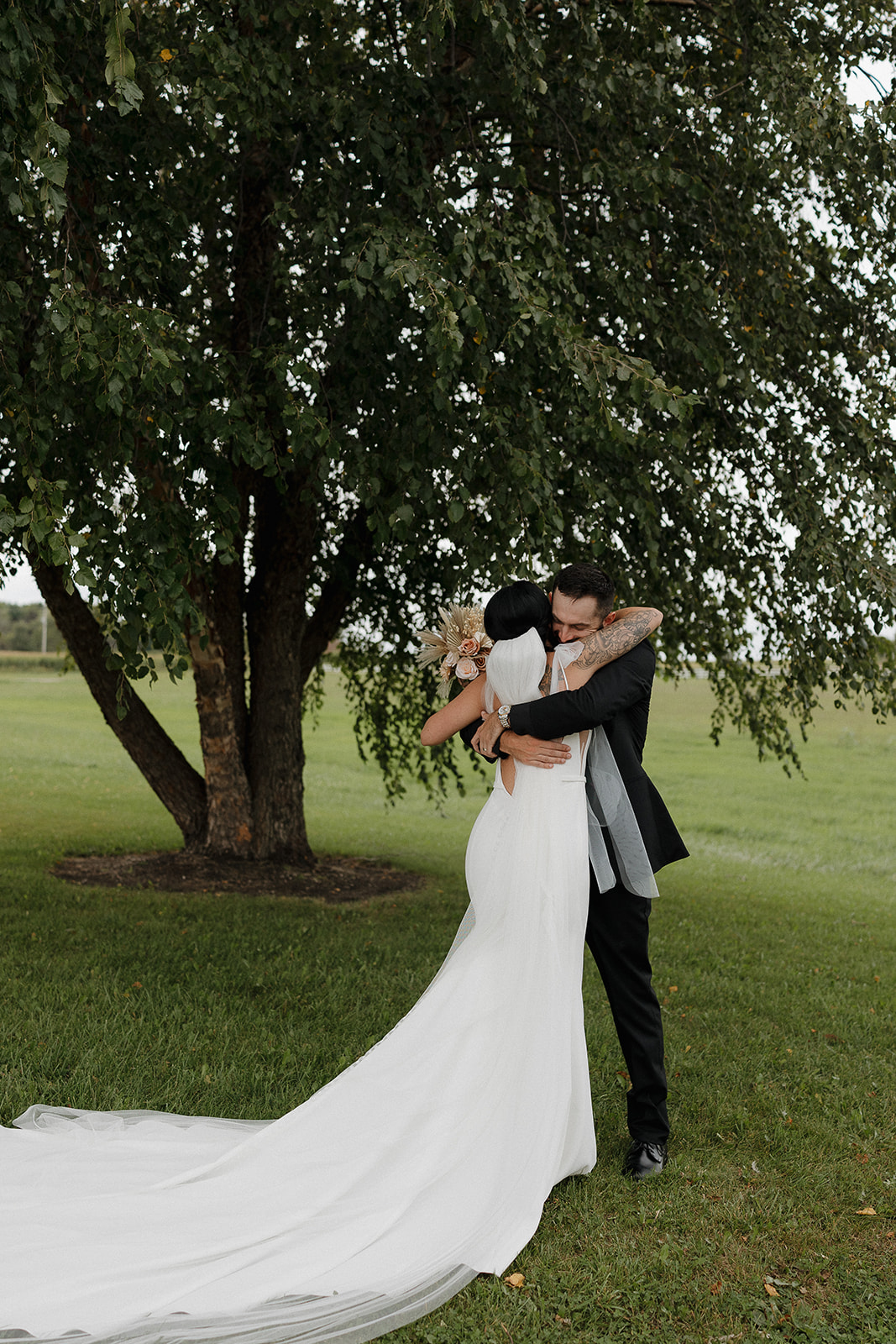 Bride and groom sharing an embrace under a tree with her veil catching the breeze