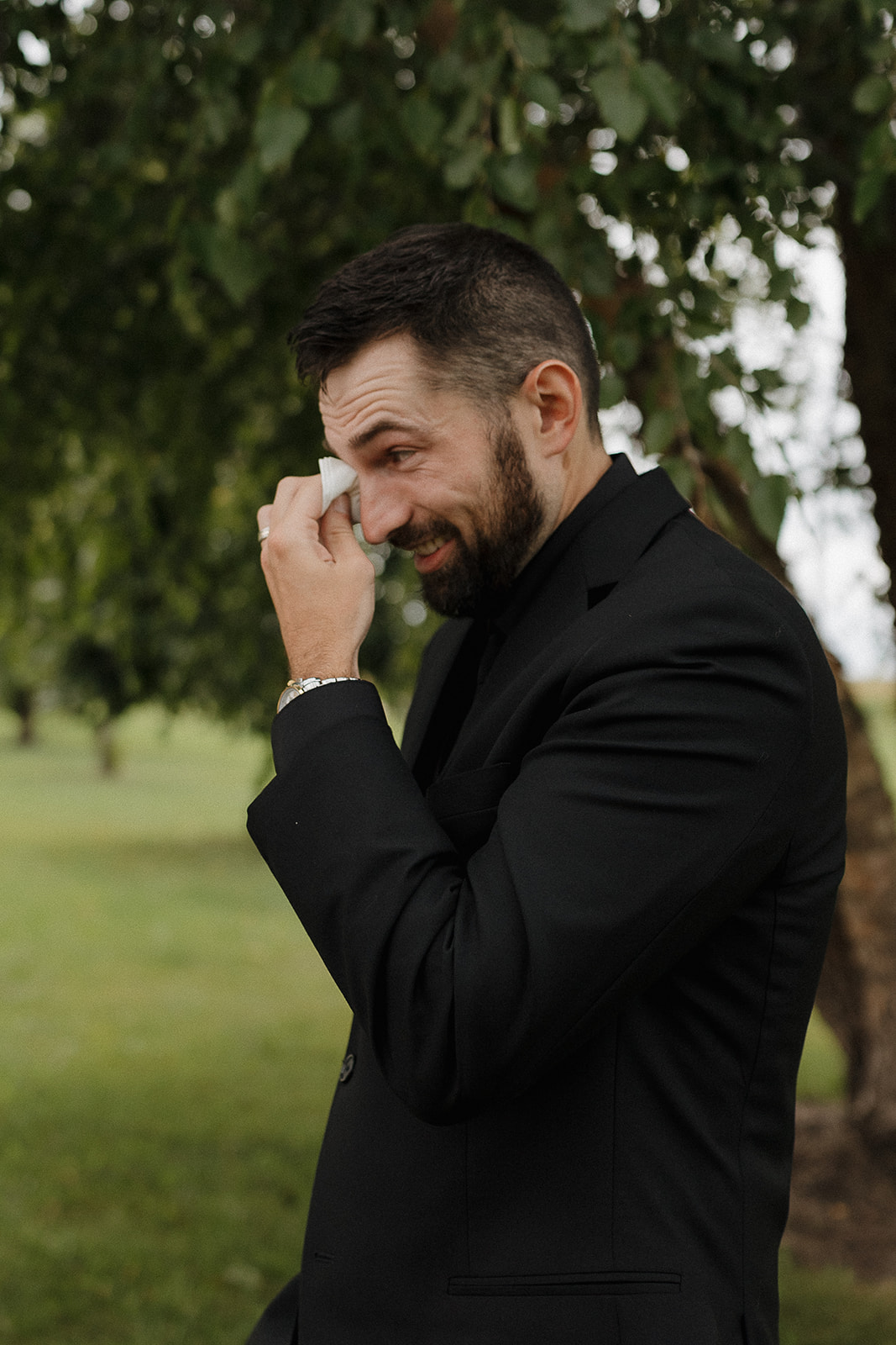 Groom wiping away happy tears in his black suit under the trees at his fall wedding